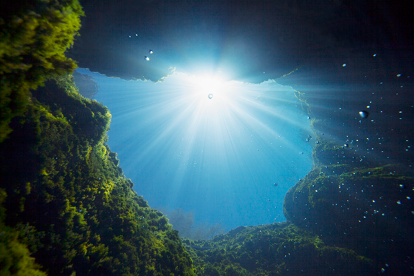 An awe-inspiring underwater view from within Jacob’s Well, a natural spring in Texas. The image is taken from deep within the cave system, looking straight up toward the surface. Sunlight pierces through the water, casting dramatic rays and illuminating the rocky, moss-covered cavern walls. Tiny air bubbles float upward, enhancing the sense of depth and serenity in this iconic geological formation known for its crystal-clear waters and vertical underwater shaft.